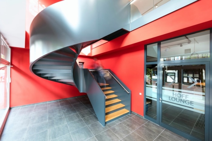 Ein modernes Interieur zeichnet sich durch eine schwarze Wendeltreppe mit Holzstufen vor leuchtend roten Wänden, grau gefliestem Boden und Glastüren mit der Aufschrift „Hoff Lounge“ aus. Eine verschwommene Gestalt steigt die Treppe hinauf.