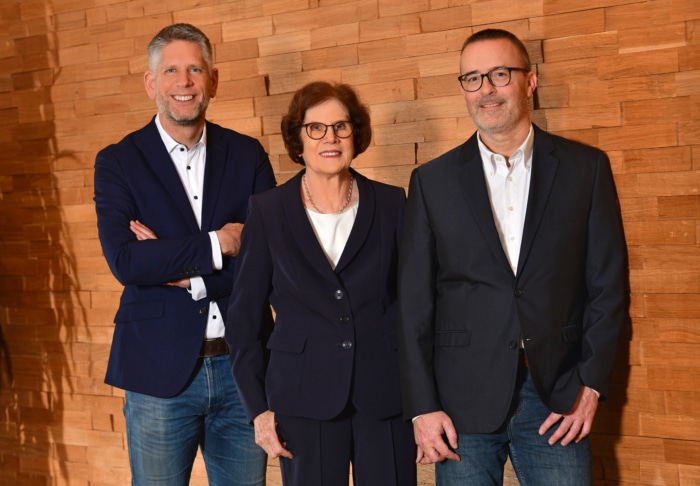 Drei Menschen stehen nebeneinander vor einer Holzwand, alle tragen dunkle Blazer und Brillen und lächeln in die Kamera – eine einladende Gruppe von Mitgliedern der Stiftung, mit den beiden Männern rechts und links der Frau in der Mitte.