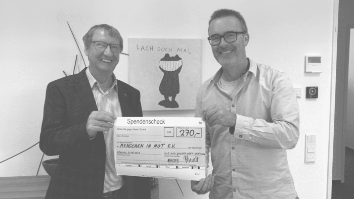 Zwei lächelnde Männer halten einen großen Spendenscheck über 270 Euro für Menschen in Not e.V., unterstützt von der Stiftung. Sie stehen im Innenbereich vor einer Wand mit einer Zeichnung und dem deutschen Spruch „Lächle doch mal“.