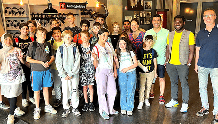 Eine bunt gemischte Gruppe von Kindern und Erwachsenen posiert bei einer Stiftungsveranstaltung gemeinsam in einem Gebäude und lächelt in die Kamera. Einige machen Peace-Zeichen. Im Hintergrund hängen Mäntel, und auf einem Schild steht „Audioguides“. Die Atmosphäre wirkt freundlich und ungezwungen.
