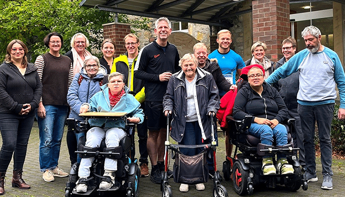 Eine Gruppe von fünfzehn Menschen, darunter Männer und Frauen mit und ohne Behinderung, lächelt gemeinsam im Freien bei einer Stiftungsveranstaltung. Zwei Frauen nutzen Elektrorollstühle, eine Frau einen Rollator. Alle wirken fröhlich und freundlich.