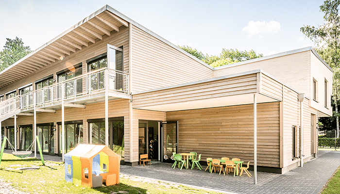 Ein modernes zweistöckiges Gebäude mit Holzfassade, großen Fenstern und einem Balkon. Draußen säumen bunte Kinderspielhäuser und Sitzgelegenheiten den gepflasterten Innenhof und erinnern an eine Kindertagesstätte oder eine Vorschule.