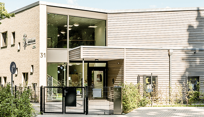 Ein modernes Stiftungsgebäude mit großen Glasfenstern, hellen Ziegel- und Holzvertäfelungen, einem schwarzen Metallzaun und Grünflächen davor. Der Eingang liegt zurückgesetzt, an der Wand prangt die Zahl 31.