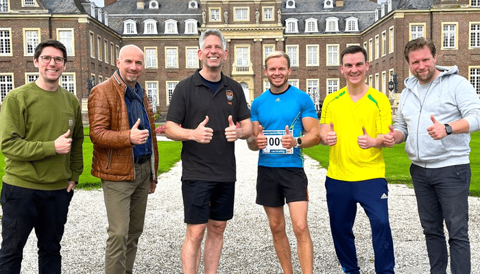 Sechs Männer stehen lächelnd und mit erhobenem Daumen vor einem großen, historischen Stiftungsgebäude. Zwei tragen Sportkleidung mit Startnummern, die anderen sind in Freizeitkleidung und Jacken auf dem Schotterweg unterwegs.